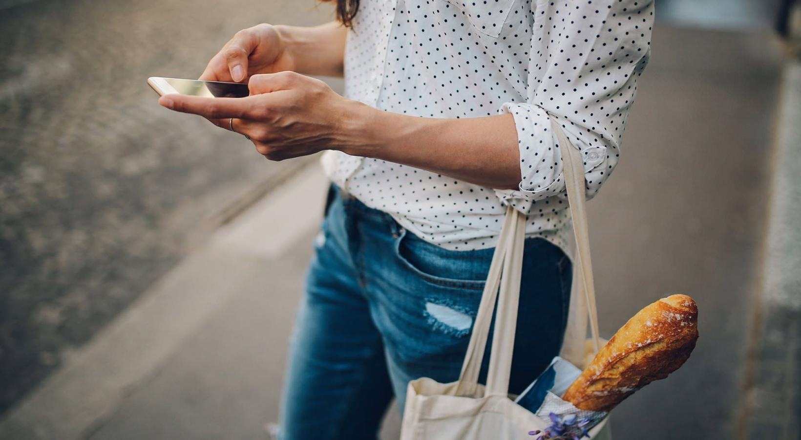 woman walks down the street and texts with a bag of groceries on her arm